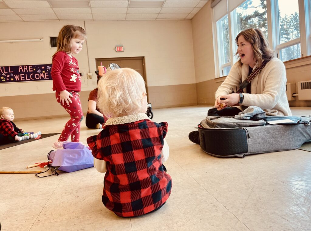 Young children attentively watching the music therapist who is seated, holding her guitar, and singing.