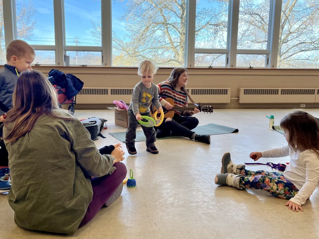 Young children accompanied by an adult sit facing the music therapist leading the class on guitar with vocals, and between them, one young boy is playing two tambourines.